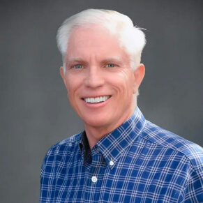 A man with short white hair in a blue plaid shirt smiles against a dark gray background.