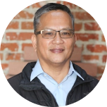 A person with glasses and a short haircut stands in front of a brick wall, smiling and wearing a collared shirt and jacket.