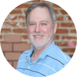 A man with gray hair and a beard smiles at the camera, wearing a light blue striped shirt in front of a brick wall.