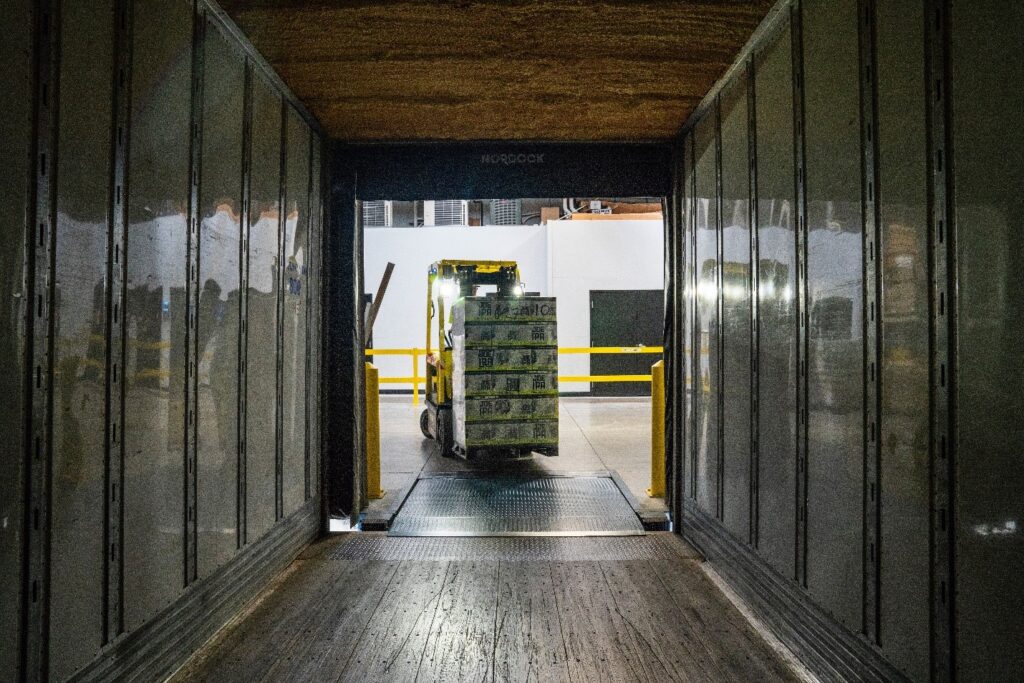 A forklift moves a stack of boxes through a narrow passage into a warehouse with yellow safety railings.