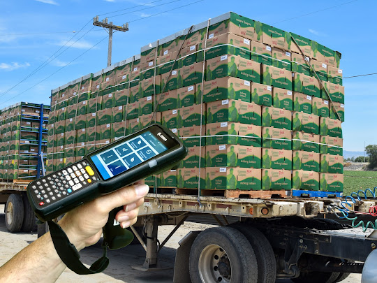 Hand holding a barcode scanner in front of a truck loaded with stacked green boxes.