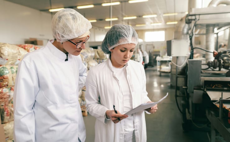 Two individuals in lab coats and hairnets inspect a document in a food processing facility.